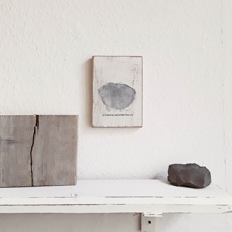 May include: A small, white wooden frame with a gray abstract painting inside. The painting is a simple, abstract shape with a rough texture. The frame is hanging on a white wall above a white shelf.