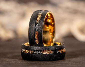 Puede incluir: Dos anillos negros con detalles dorados y plateados. Los anillos tienen un exterior negro texturizado y un interior dorado. Los anillos están apilados, destacando el diseño detallado y la artesanía.
