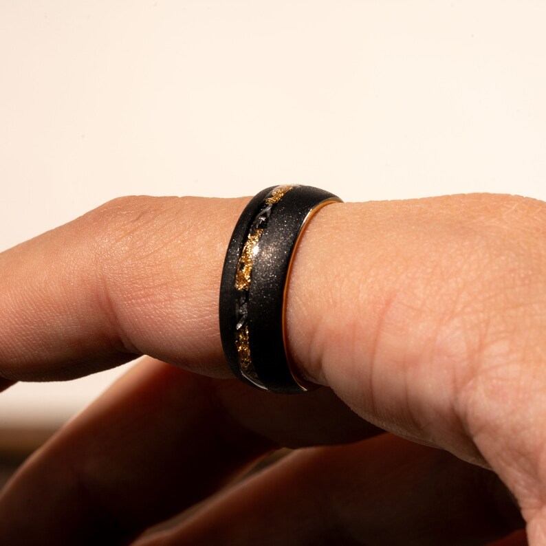 May include: A black ring with gold detailing. The ring has a black, textured band with gold-coloured flecks and a gold inner band. The ring is displayed on a finger. The ring's design creates a striking contrast.