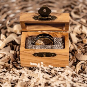 May include: An open wooden ring box with a brass clasp displays a black and gold ring. The ring features a black band with gold and silver detailing. The box is lined with hessian and rests on wood shavings.