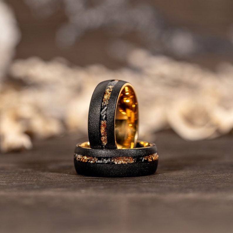 May include: Two black rings with gold and silver detailing. The rings feature a textured black exterior and a gold interior. The rings are stacked, highlighting the detailed design and craftsmanship.