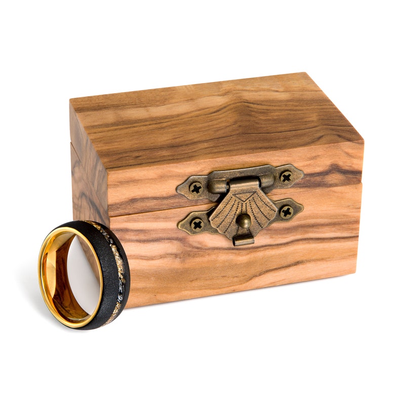 May include: A black and gold ring with a textured band and a gold interior sits beside a wooden box with a brass clasp. The ring has a black band with a gold interior and a textured surface.