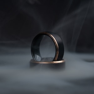 May include: Two black rings with a gold-colored band. The rings are set against a smoky, dark background. The rings are likely wedding bands or fashion accessories.