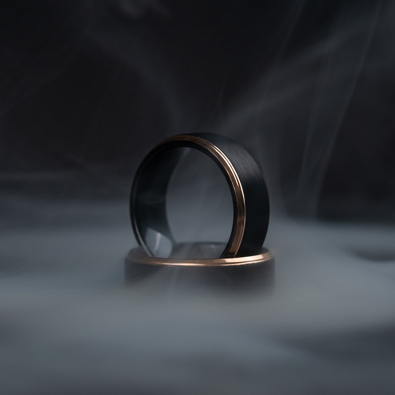 May include: Two black rings with a gold-colored band. The rings are set against a smoky, dark background. The rings are likely wedding bands or fashion accessories.