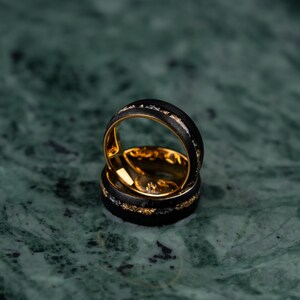 May include: Two black rings with gold interiors and accents. The rings feature a textured black band with gold and silver flecks. The rings are stacked, highlighting their design and craftsmanship.