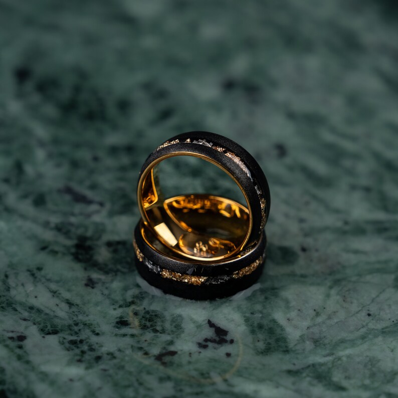 May include: Two black rings with gold interiors and accents. The rings feature a textured black band with gold and silver flecks. The rings are stacked, highlighting their design and craftsmanship.