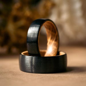 May include: Two wedding bands, one with a black brushed finish and the other with a wood inlay. The bands are stacked on top of each other.