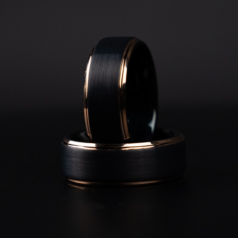 May include: Two black rings with a brushed metal finish and a gold-colored inner and outer rim. The rings are stacked on top of each other against a dark background, showcasing their contrasting textures and colors.
