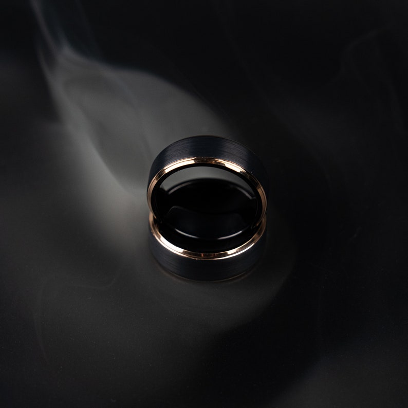 May include: A black ring with a brushed metal finish and a gold-colored inner band. The ring is reflected on a dark surface, with a wisp of white smoke in the background. The ring is a close-up shot.