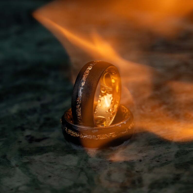 May include: Two black rings with gold detailing are stacked. The rings have a dark exterior and a gold interior. Gold accents are visible as a band around the outside and flecks. The background is a blurred orange and green, creating a warm glow.