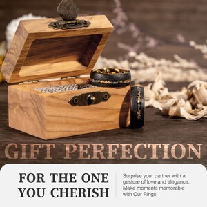 May include: A wooden ring box with an open lid displays two black rings with gold accents. The rings have a textured appearance. The box is made of light brown wood and has a metal clasp. The text "GIFT PERFECTION" is visible.