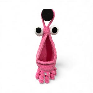 May include: A pink crocheted hanging basket shaped like a cartoon squid with large white and black eyes. The basket has a round black hanging loop and several curled tentacles at the bottom.