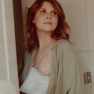 May include: A woman with red hair is wearing a light green robe with lace trim. She is wearing a white mesh bra underneath the robe. She has a small tattoo on her shoulder.