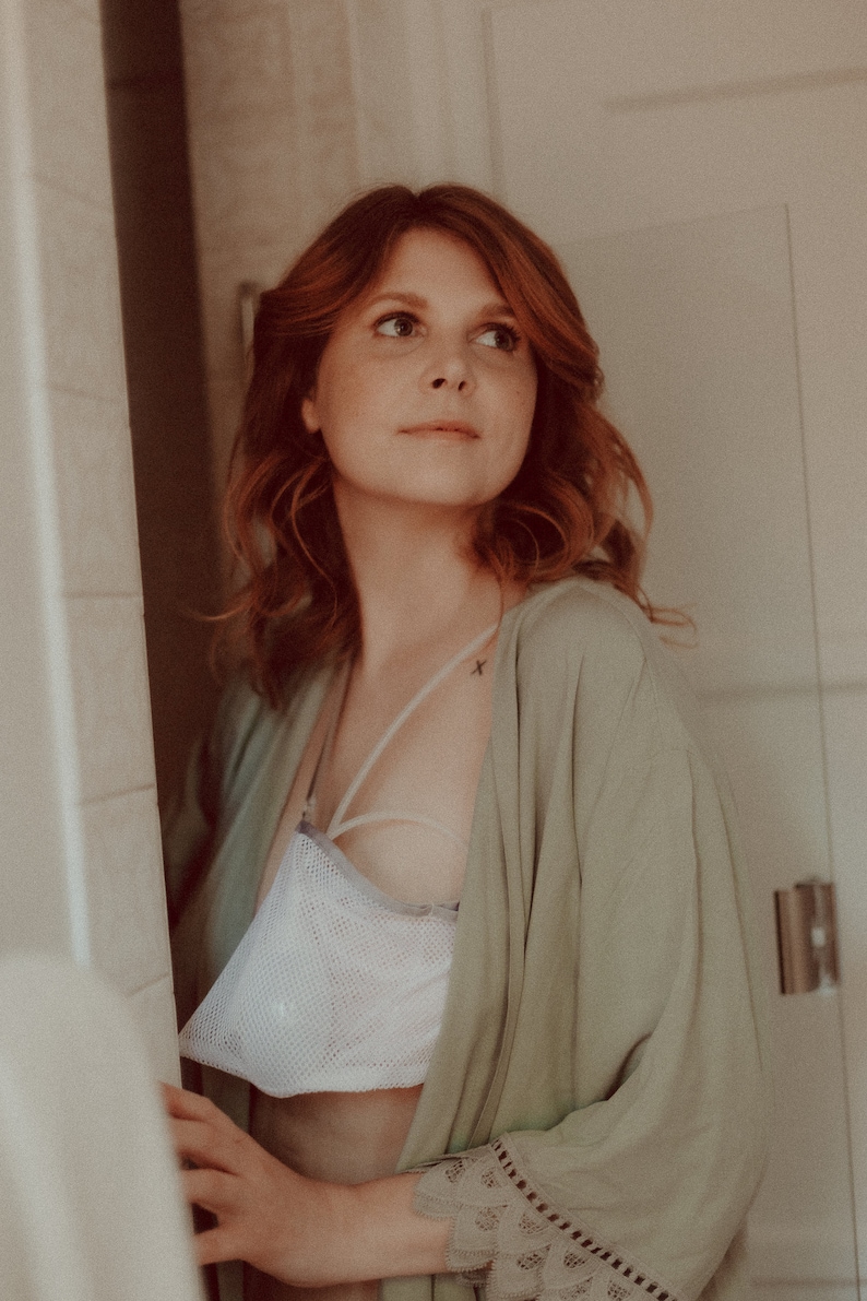 May include: A woman with red hair is wearing a light green robe with lace trim. She is wearing a white mesh bra underneath the robe. She has a small tattoo on her shoulder.