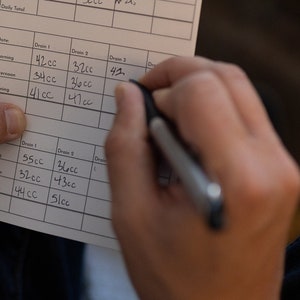 May include: A close-up of a notebook with handwritten entries in a table format. The table has columns labeled "Date", "Drain 1", "Drain 2", and "Drain 3". The entries in the table are in milliliters (cc).