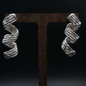 Puede incluir: Un par de pendientes de alambre de plata con un diseño tejido. Los pendientes se muestran en un soporte de madera.