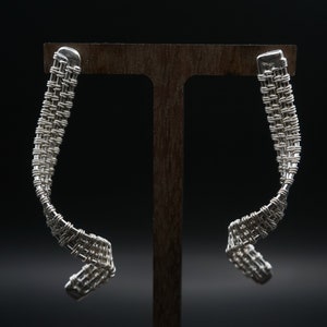 Puede incluir: Un par de pendientes de alambre de plata con un diseño tejido. Los pendientes se muestran en un soporte de madera.