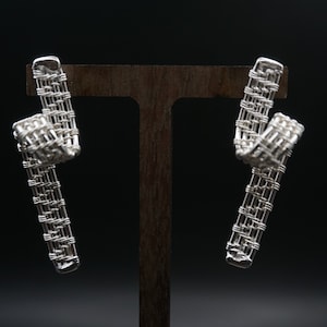 Puede incluir: Un par de pendientes de plata con un diseño tejido. Los pendientes se muestran en un soporte de madera.