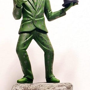 May include: A figurine of a character in a green suit covered in question marks, a purple mask, and a bowler hat. The figure is standing on a stone-like base. The character is posed with one hand raised and the other holding the hat.