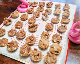 May include: A white baking sheet with a brown, circular, patterned surface. The sheet is filled with pecan pralines, which are small, round, and have a light brown color. The pralines are arranged in rows and columns on the sheet.
