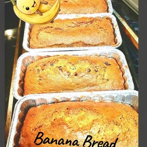 May include: Four loaves of freshly baked banana bread in aluminum foil pans. The loaves are golden brown with a slightly textured surface. The words "Banana Bread" are written in a cursive font on the bottom loaf. A cartoon banana is in the upper left corner.