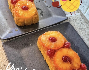 May include: Two rectangular slices of upside-down cake on dark gray plates. Each cake is topped with pineapple rings and cherries. A cartoon pineapple wearing sunglasses is in the background. The text "Upside Down" is written on the plate.