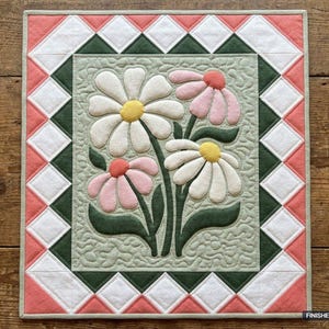 May include: A rectangular quilt featuring a central panel with appliqued white and pink daisies on a sage green background. The border consists of a geometric diamond pattern in white, dark green, and coral pink. Finished size 91.44 cm x 106.68 cm.