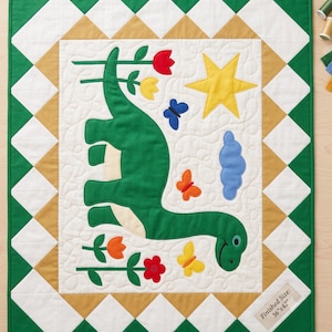 May include: A rectangular quilt featuring a large green dinosaur appliqué on a white background with swirling stitching. The design includes a yellow star, a blue cloud, colorful butterflies, and flowers. It has a diamond-patterned border in green, white, and tan, with a label stating a finished size of 36 inches by 42 inches.