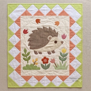 Könnte beinhalten: Ein gesteppter Wandbehang mit einem cremefarbenen Mittelteil, auf dem ein brauner Igel appliziert ist, umgeben von Stoffblumen und Blättern. Der Rand besteht aus weißen und pfirsichfarbenen Dreiecken mit einem lindgrünen Saum. Fertiggröße 91cm x 107cm.