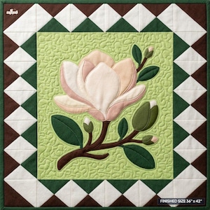 Könnte beinhalten: Ein quadratischer, gesteppter Wandbehang mit einem hellgrünen Mittelteil, auf dem eine große applizierte Magnolienblüte in zartem Rosa und Creme, dunkelgrüne Blätter und braune Stiele zu sehen sind. Der Rand zeigt ein geometrisches Muster aus weißen und braunen Dreiecken. Fertige Größe 91,44 cm x 106,68 cm.