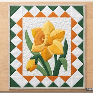 Puede incluir: Un tapiz de tela rectangular con una aplicación central de narciso amarillo sobre un fondo acolchado blanco. El borde presenta un patrón geométrico de triángulos verdes, blancos y naranjas. El texto indica: FINISHED SIZE 91 cm x 107 cm.