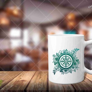 Peut inclure: Une tasse en c&eacute;ramique blanche avec un motif de boussole bleu sarcelle entour&eacute; de fleurs. La boussole a quatre points et est entour&eacute;e d'une couronne de fleurs.