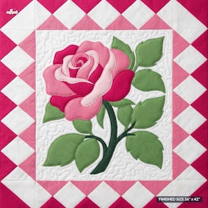 Op de afbeelding: Een vierkant quiltblok met een centrale applicatie van een roze roos met groene bladeren op een witte doorgestikte achtergrond. De rand bestaat uit een ruitpatroon in afwisselende tinten roze en wit. Afgewerkte maat 91,44 cm bij 106,68 cm.