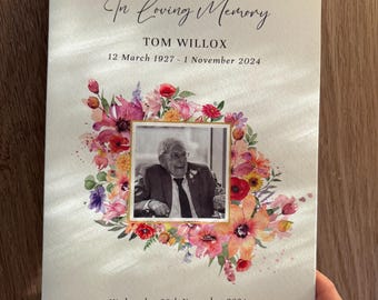 May include: A memorial booklet with the text "In Loving Memory" and the name "TOM WILLOX" with dates. A photo is surrounded by a floral arrangement. The booklet is held by a person.