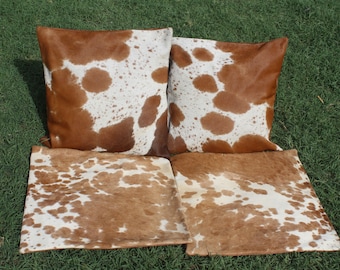 May include: Four brown and white cowhide throw pillows with a spotted pattern. The pillows are arranged in a row on a green grassy surface.
