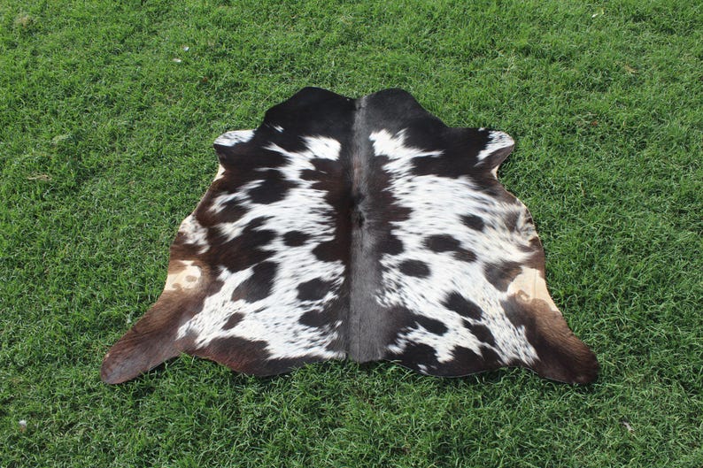 Puede incluir: Una alfombra de piel de vaca negra y blanca con una franja central marr&oacute;n. La alfombra tiene un aspecto natural y r&uacute;stico y es perfecta para a&ntilde;adir un toque de estilo r&uacute;stico a cualquier habitaci&oacute;n.