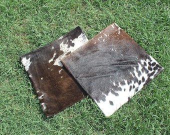 May include: Two cowhide throw pillows with a black and white pattern. The pillows are on a green grassy surface.