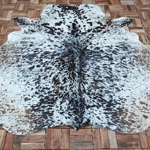 May include: A cowhide rug with a natural spotted pattern in black, white, and brown. The rug is laid out on a wooden deck, showcasing its irregular shape and texture. The hide's unique markings create a striking visual contrast.