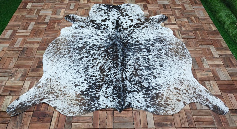 May include: A cowhide rug with a natural spotted pattern in black, white, and brown. The rug is laid out on a wooden deck, showcasing its irregular shape and texture. The hide's unique markings create a striking visual contrast.