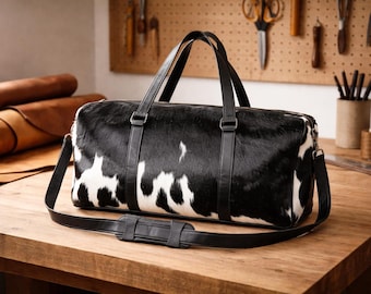 May include: A duffel bag featuring a black and white cowhide pattern with black leather trim, two top handles, and a detachable shoulder strap with a padded section. The bag has a rectangular shape with rounded edges and a top zipper closure.