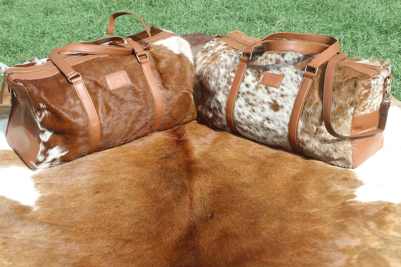 May include: Two brown and white cowhide leather duffel bags with brown leather straps and handles. The bags are sitting on a brown and white cowhide rug.