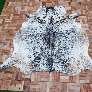 May include: A cowhide rug with a natural spotted pattern in black and brown on a white background. The rug is irregularly shaped, with a smooth texture. The rug is displayed on a wooden tiled surface.