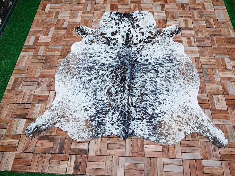 May include: A cowhide rug with a natural spotted pattern in black and brown on a white background. The rug is irregularly shaped, with a smooth texture. The rug is displayed on a wooden tiled surface.