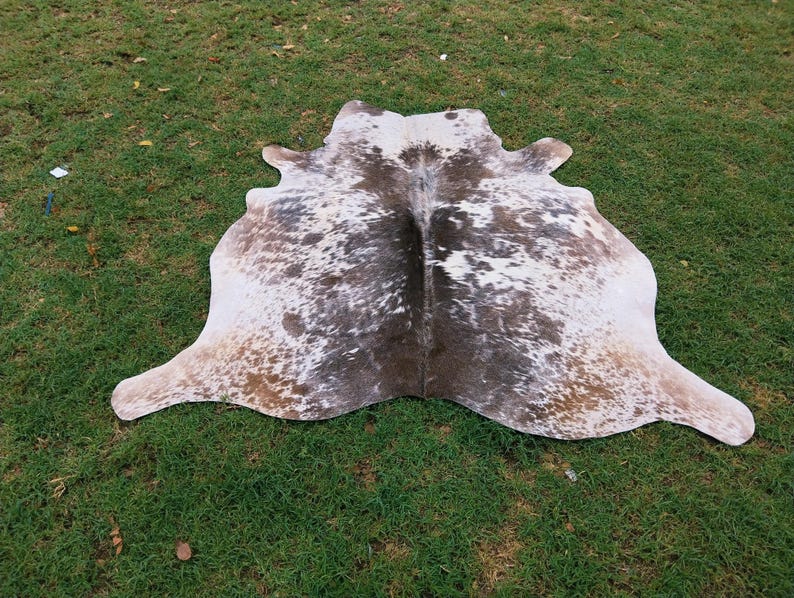 May include: A cowhide rug with a natural pattern of white, brown, and tan spots. The rug is irregularly shaped, with rounded edges and a soft texture. It is lying on a bed of green grass.