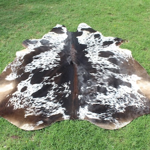 Puede incluir: Una alfombra de piel de vaca marr&oacute;n y blanca con un patr&oacute;n de manchas. La alfombra est&aacute; colocada sobre una superficie de hierba verde.