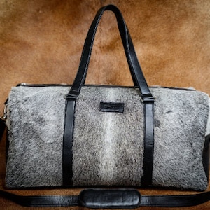 May include: A black leather duffel bag with a grey and white cowhide pattern. The bag has a top handle and a shoulder strap.