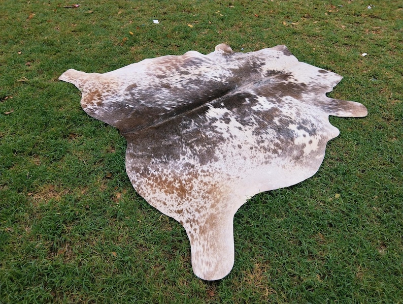 May include: A cowhide rug with a natural pattern of white, brown, and tan spots. The rug is irregularly shaped and lies on green grass. The hide has a soft, textured appearance.
