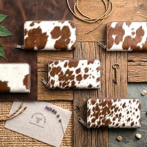 May include: Five rectangular wallets featuring a brown and white cowhide pattern with a textured hair-on-hide finish. Each wallet is secured with a silver-toned metal zip closure along the top and side edges, showcasing a consistent spotted design.