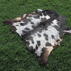 Puede incluir: Una alfombra de piel de vaca negra y blanca con un patr&oacute;n de manchas. La alfombra est&aacute; colocada sobre una superficie de hierba verde.
