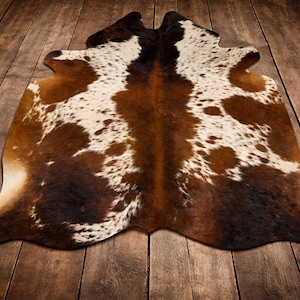 May include: A cowhide rug featuring a natural, irregular shape with a tri-colour pattern of dark brown, light tan, and white patches. The hide displays a dense, short-haired texture with organic, asymmetrical markings across the entire surface.